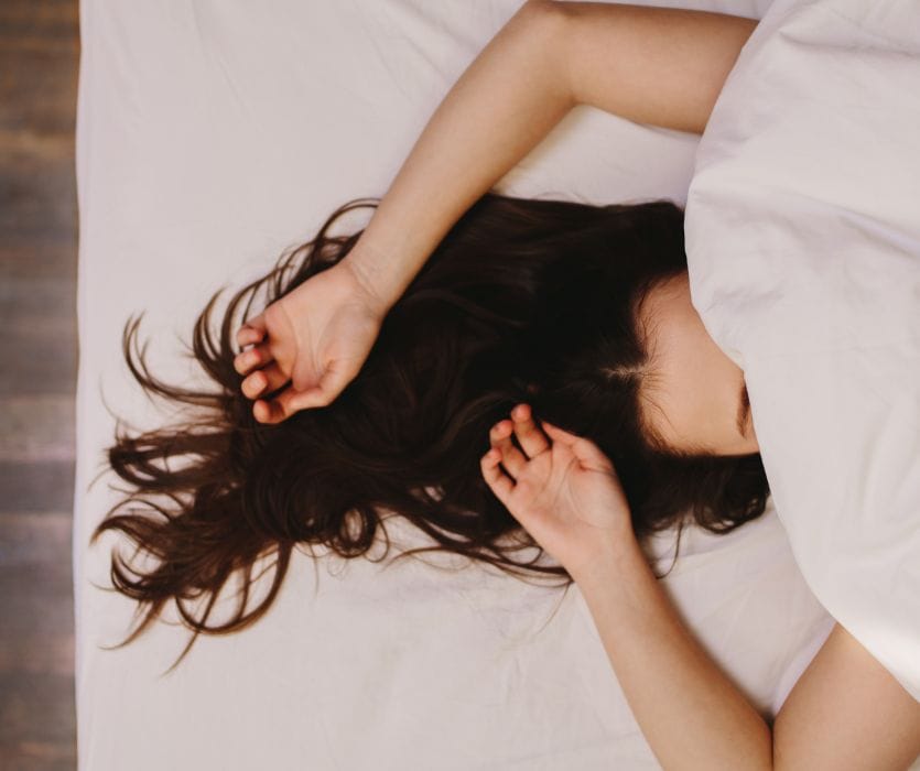Pregnant woman lies on her back in bed with arms above her head, covered by white bedding, reflecting the question of sleeping on your back while pregnant.