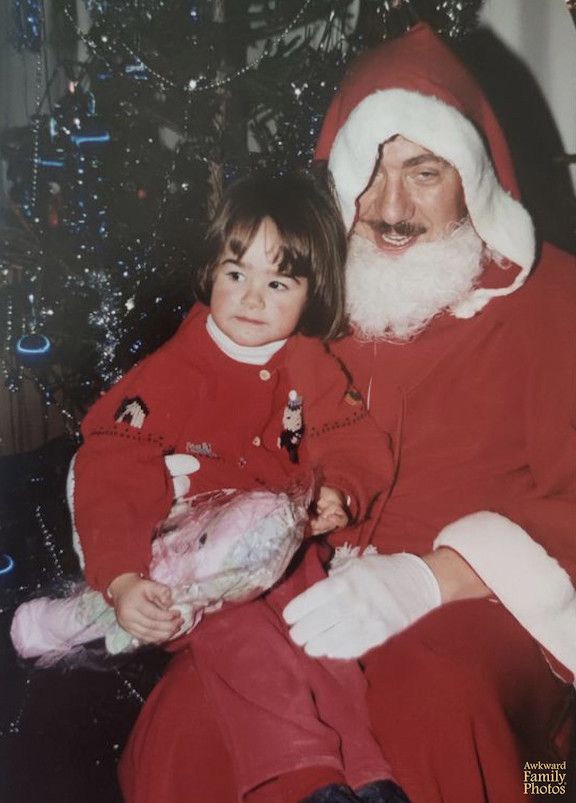 A young girl sits on the lap of a man in a Santa suit with a crooked hat and fake beard in front of a Christmas tree.