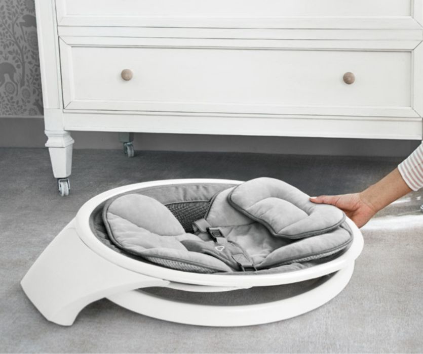 A person slides a folded, grey and white baby bouncer underneath a white dresser for compact storage.