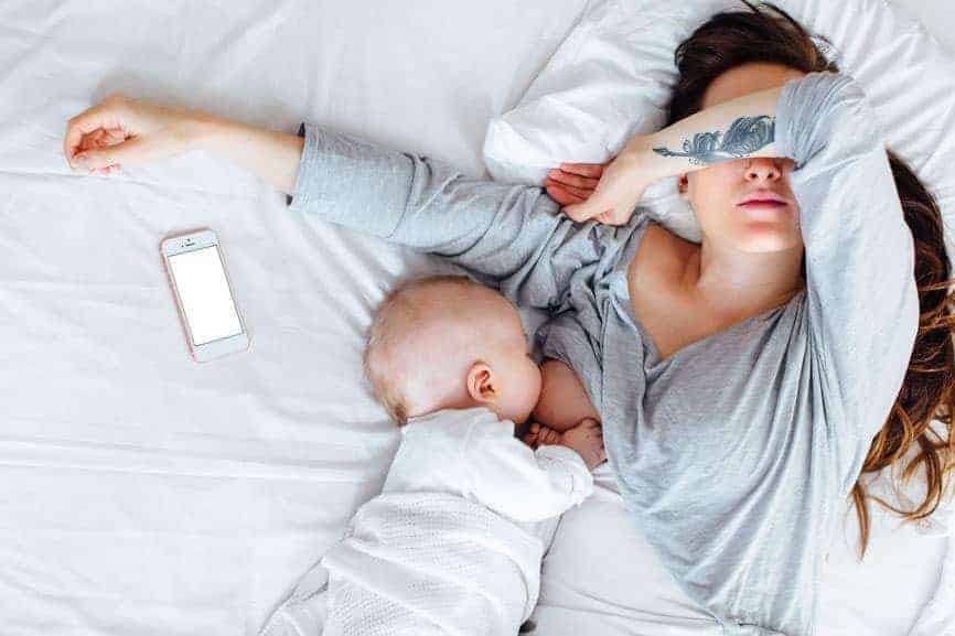 Exhausted woman in bed breastfeeding a baby with an arm covering her eyes and a smartphone lying nearby.