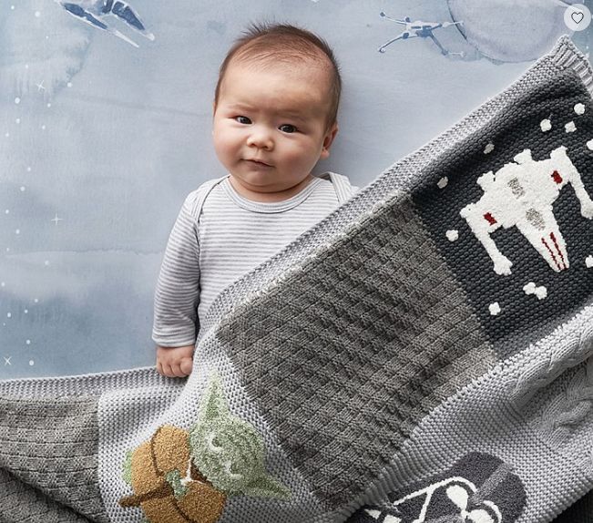 A baby lying on a Star Wars themed backdrop with a grey knit blanket featuring Yoda and an X-wing fighter.