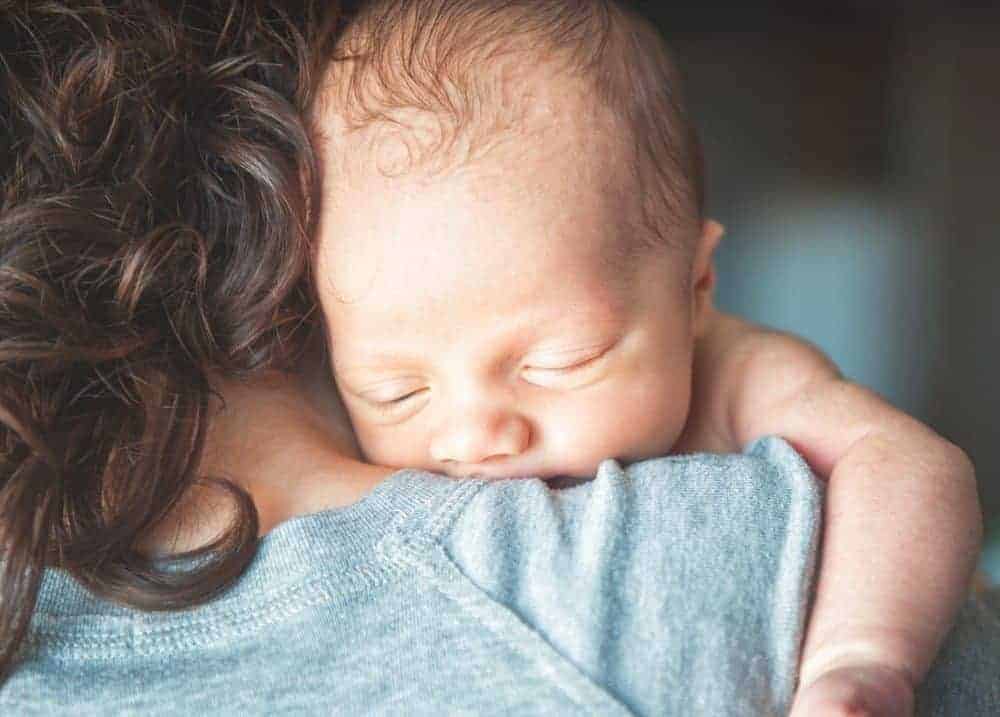 A sleeping newborn infant rests their head on a person's shoulder.