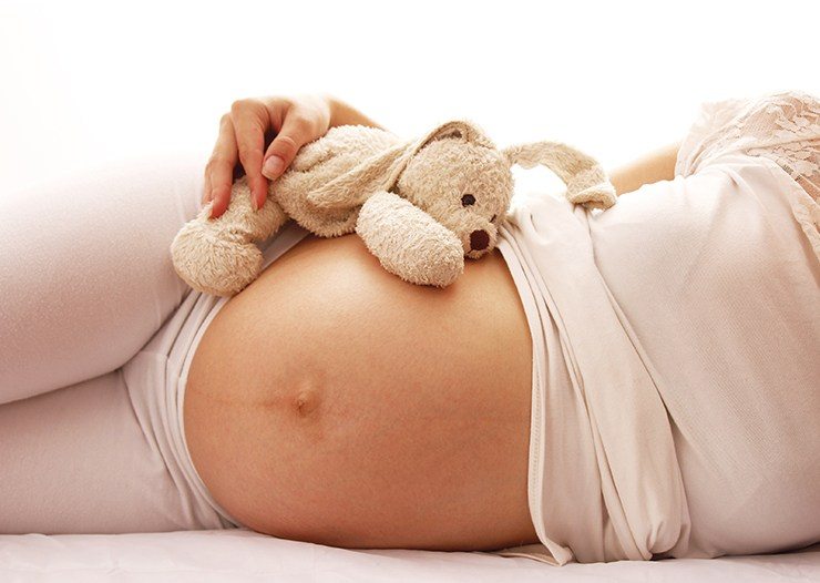 Close-up of a pregnant person's bare stomach as they lie down, with a plush stuffed rabbit resting on top.