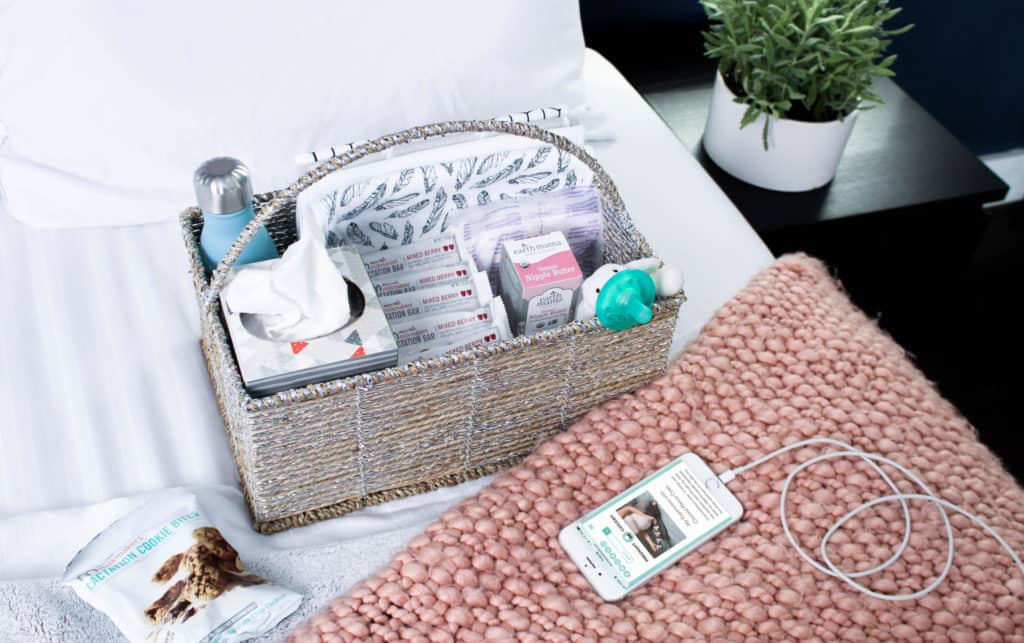 Woven bedside basket holding tissues, snacks, and baby supplies next to a charging phone on a pink knit blanket.