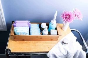 Postpartum care kit in a wooden bin with pads, peri bottle, medications, and flowers on a rolling cart.