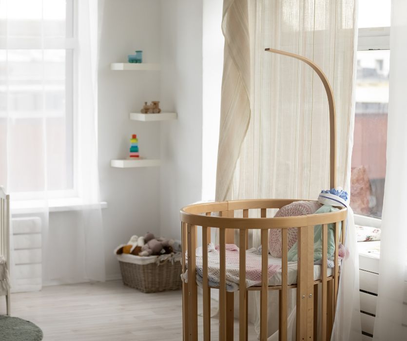 Empty wooden bassinet in a bright, white nursery next to a window with sheer curtains.