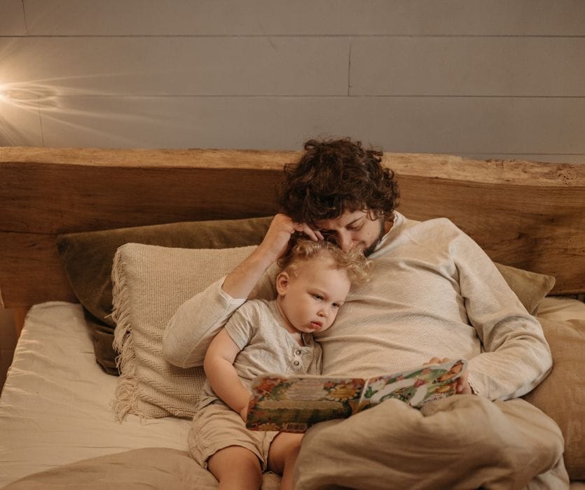 A father and young child sit together on a bed reading an illustrated storybook.