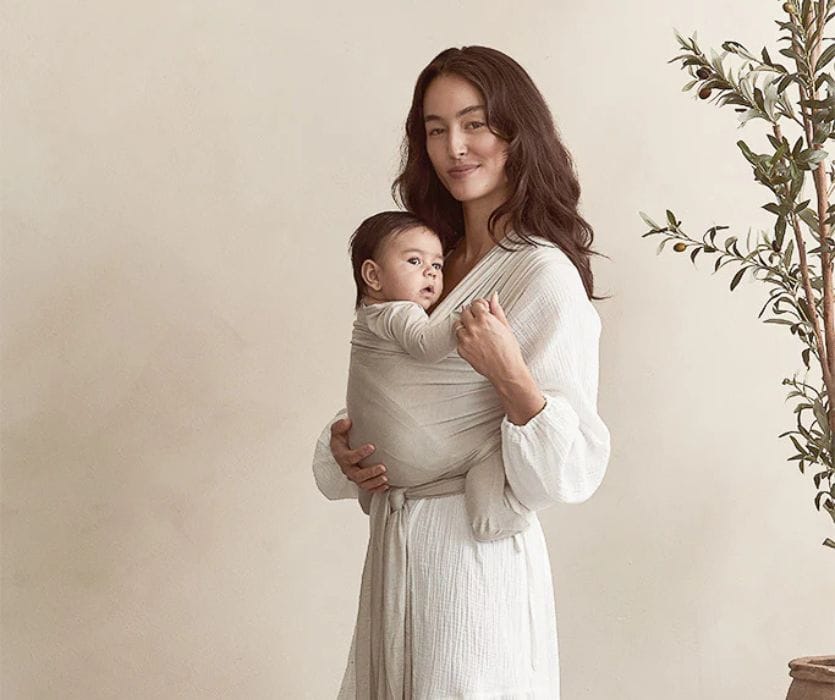 Woman wearing a baby in a neutral-toned stretchy wrap carrier.