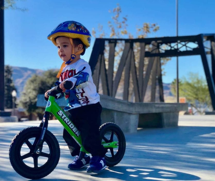 Toddler wearing a blue helmet and elbow pads riding a green Strider balance bike at a park.