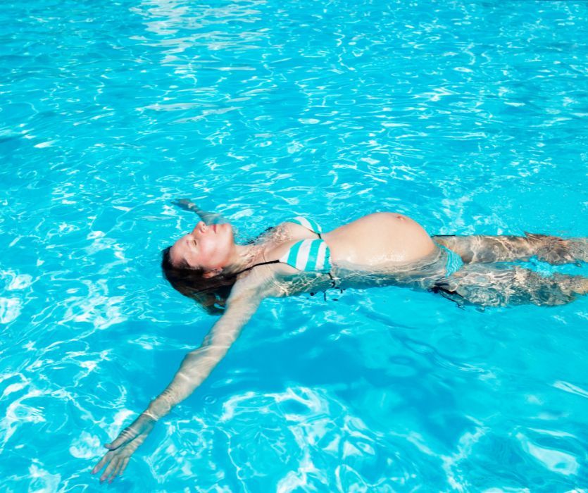 Pregnant woman in a bikini floating on her back in a turquoise swimming pool under bright sunlight.