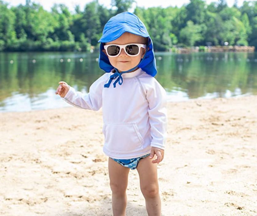 Toddler on a beach wearing a blue sun hat with a neck flap, white sunglasses, and a long-sleeve swim shirt.
