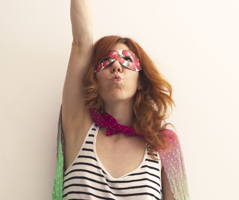 Red-haired woman in a strawberry mask and cape raising one arm in a superhero pose.