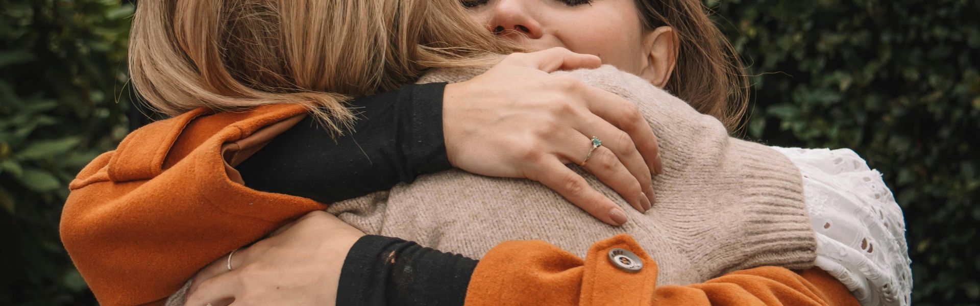 Close-up of two people embracing outdoors. One person wears an orange coat and the other a beige sweater.