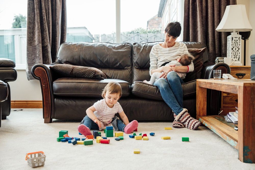 A toddler plays with colorful blocks on the floor while a woman sits on a sofa nearby holding a sleeping baby.