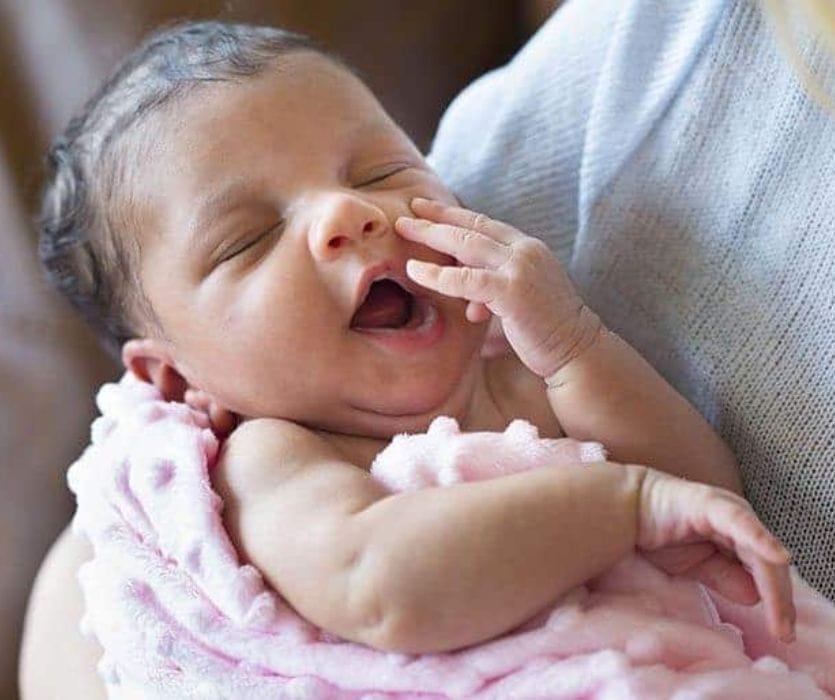 Newborn baby with eyes closed yawning while being held in a pink blanket.