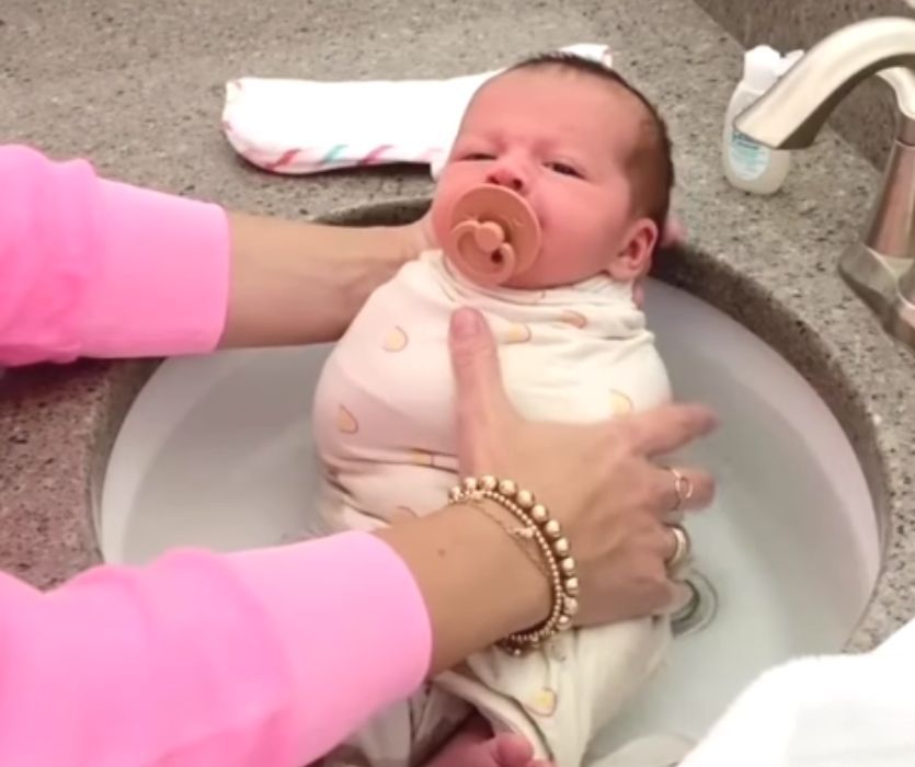 Hands holding a swaddled infant with a pacifier during a bath in a bathroom sink.