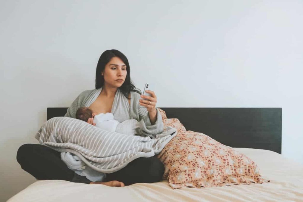 A woman breastfeeding her infant on a bed while looking at a smartphone held in her hand.