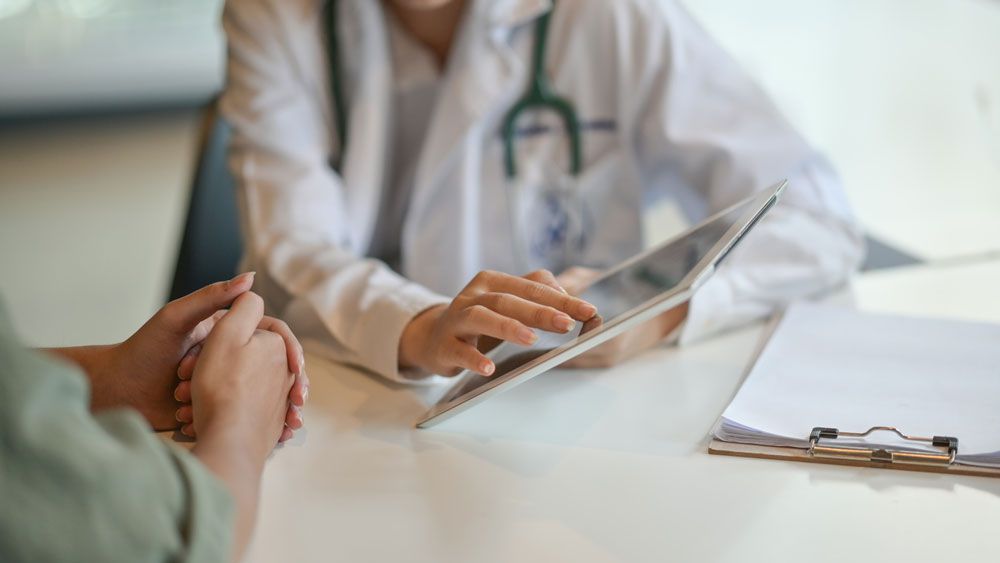 A doctor in a white coat shows information on a digital tablet to a patient during a consultation.