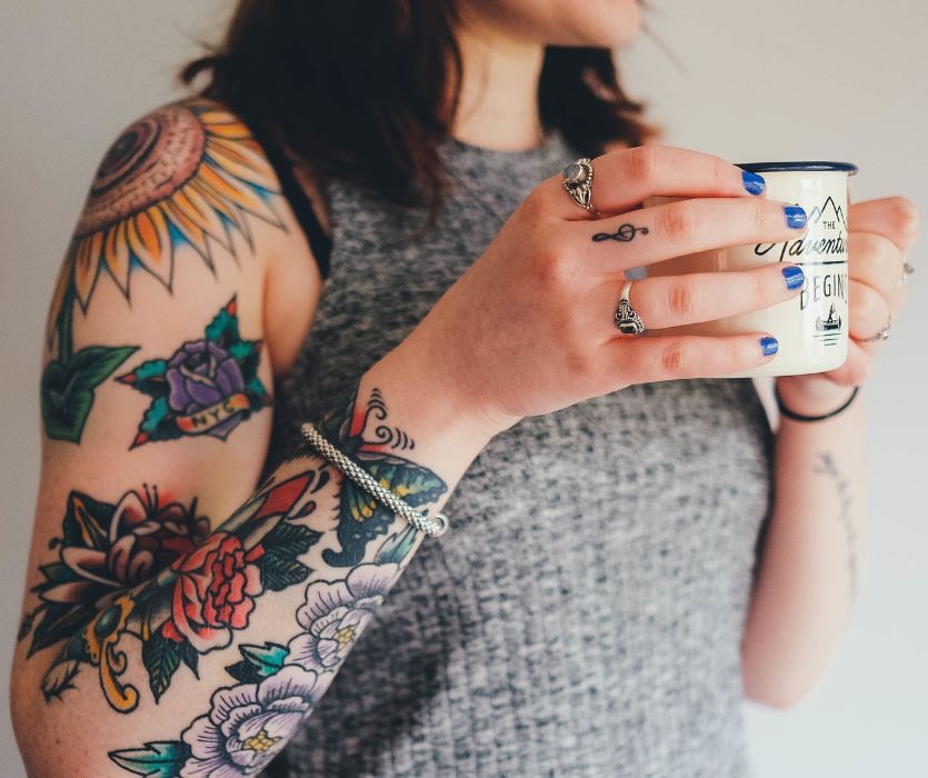 A person with colorful floral arm tattoos and small finger tattoos holding a white mug.
