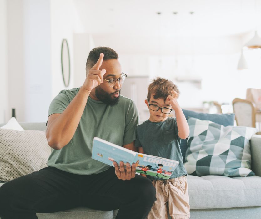 A father and young child wearing glasses practice baby sign language together while sitting on a bright couch with a book open between them at home.