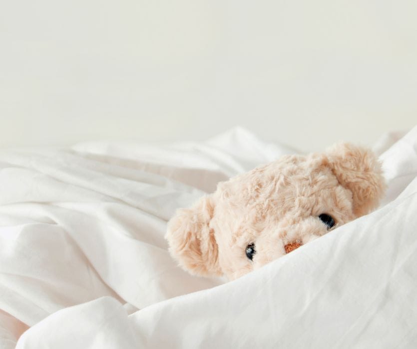 A tan fluffy teddy bear peeking out from under white bed sheets.