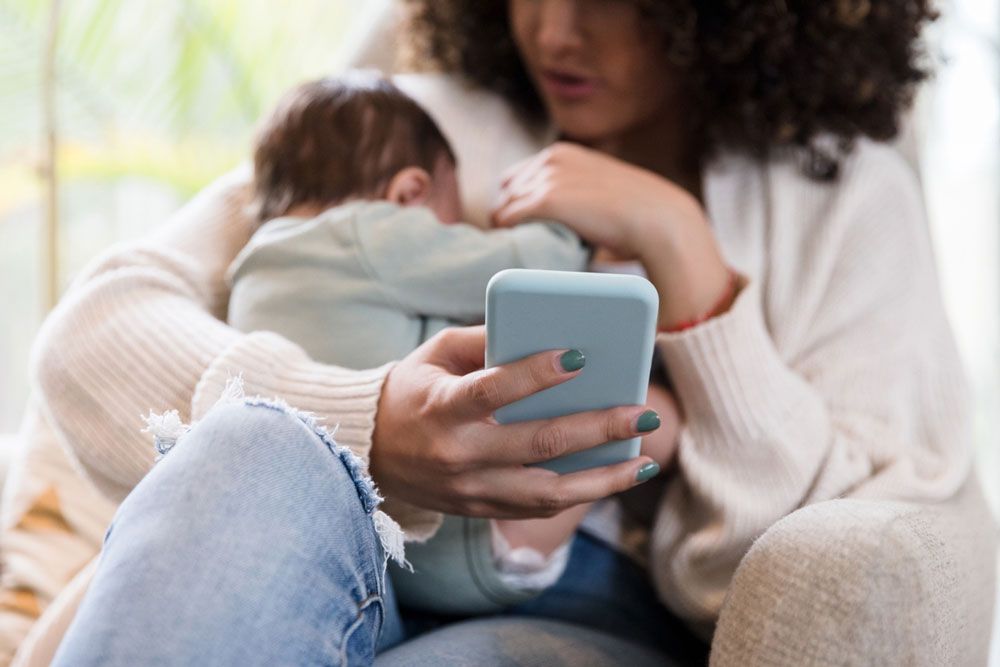 A mother holds a smartphone in one hand while cradling her infant during breastfeeding.