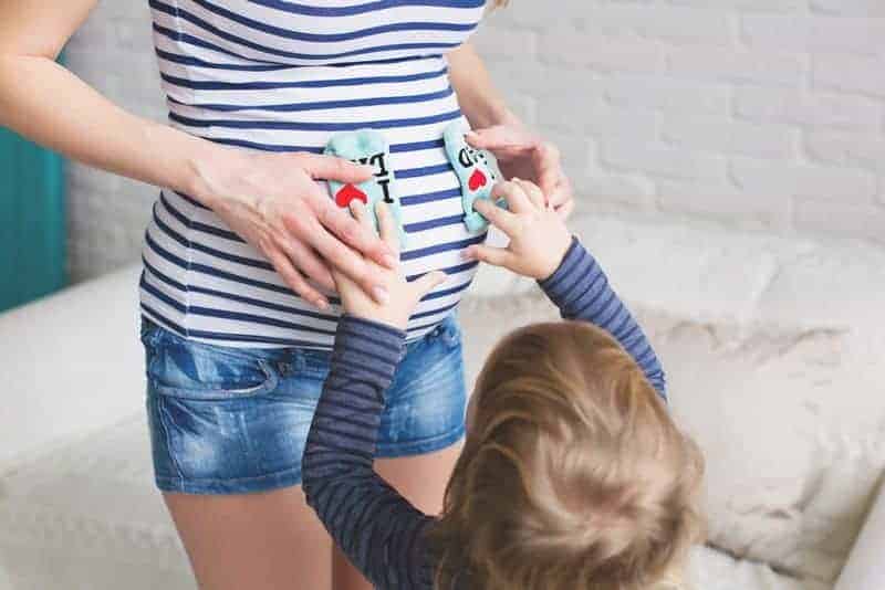 A pregnant woman and a young child hold a pair of baby socks against her baby bump.