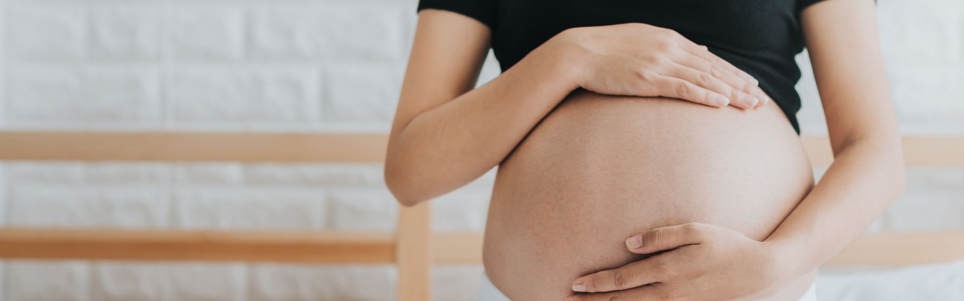 A pregnant person gently cradles their bare belly with both hands while wearing a black top in a bright room.