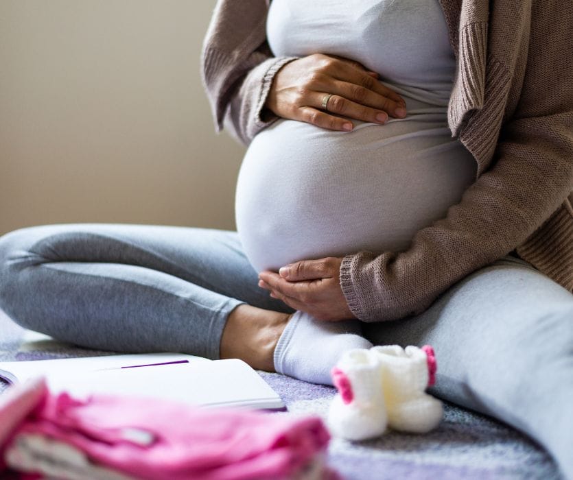 A pregnant person sitting cross-legged while holding their belly, with baby clothes and a notebook nearby.