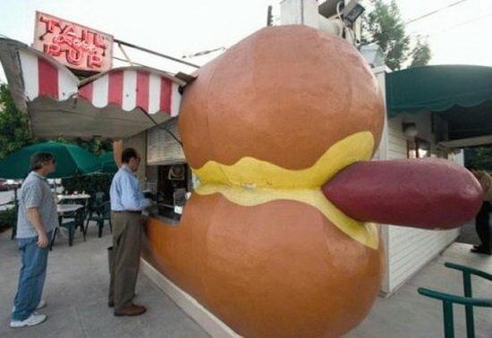 A giant hot dog sculpture sits outside a stand, with a mustard-colored filling between oversized bun halves under a striped awning.
