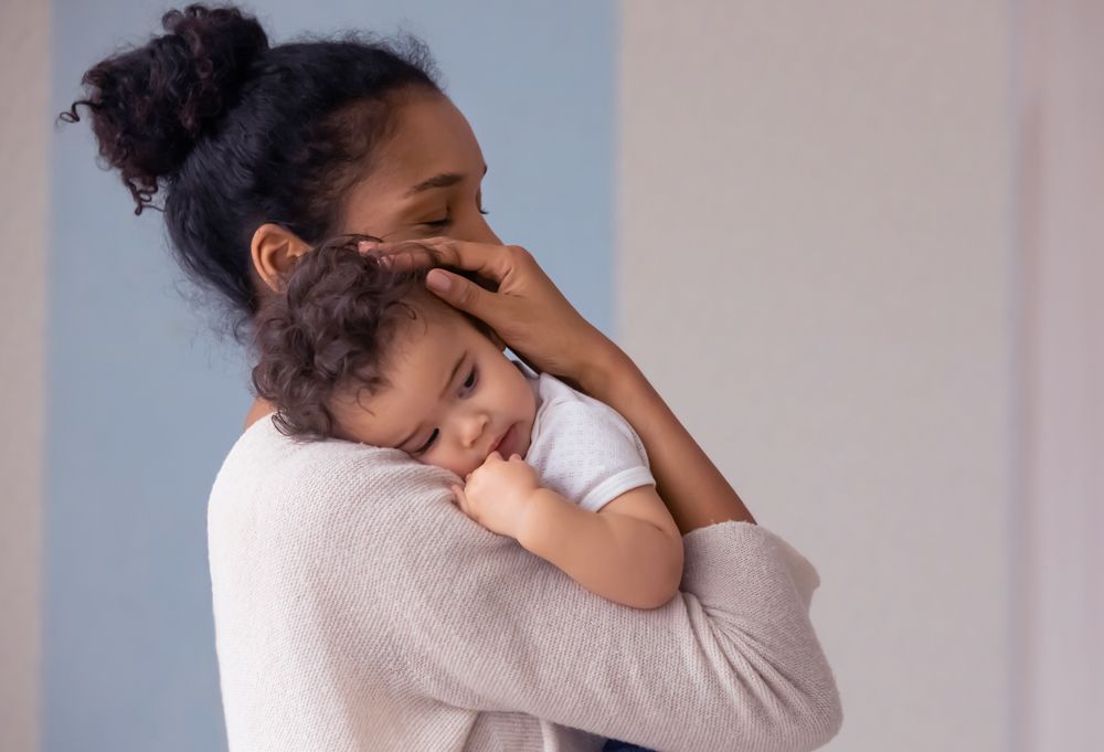 A woman holds a curly-haired baby over her shoulder while gently touching the infant's head.