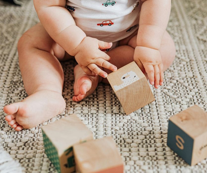 Baby sitting on a textured rug playing with wooden alphabet blocks.
