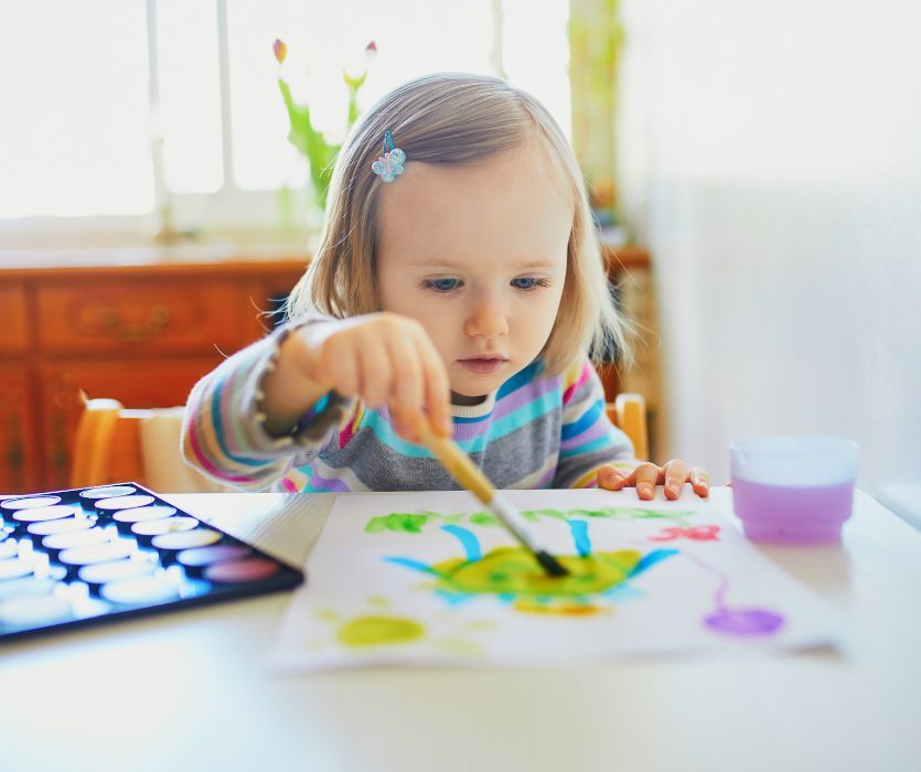 A toddler girl concentrates on painting with watercolors on a piece of white paper at a table.