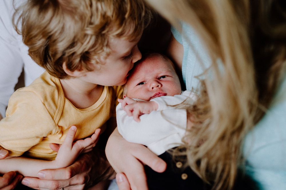 A young boy kisses the forehead of a newborn baby held in an adult's arms.