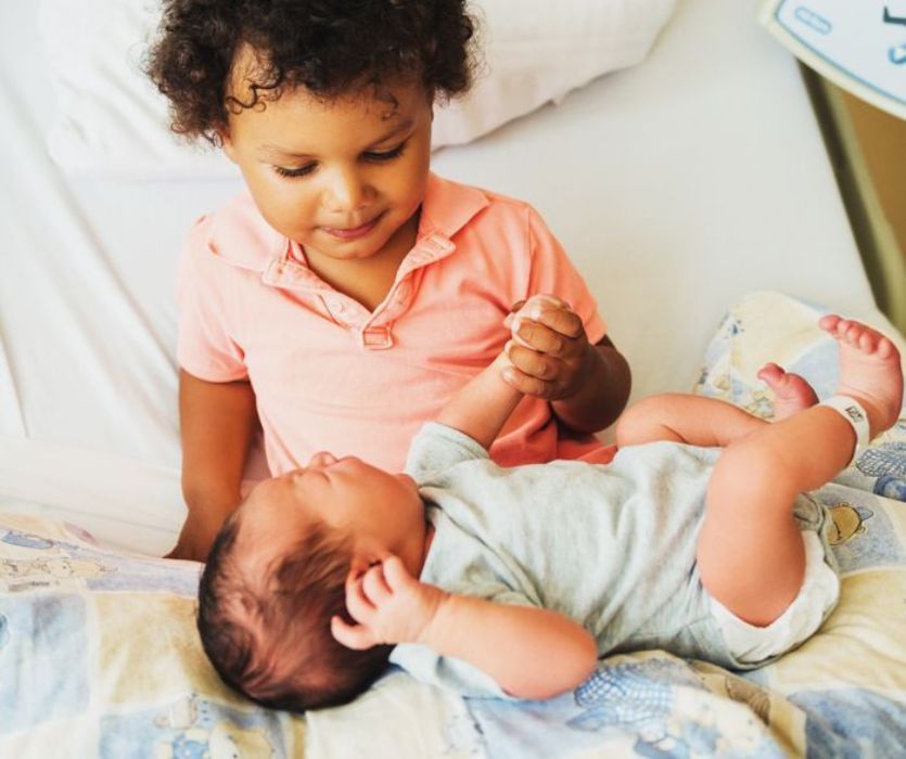 A young boy in a pink shirt sits on a hospital bed and gently holds the hand of a newborn baby.