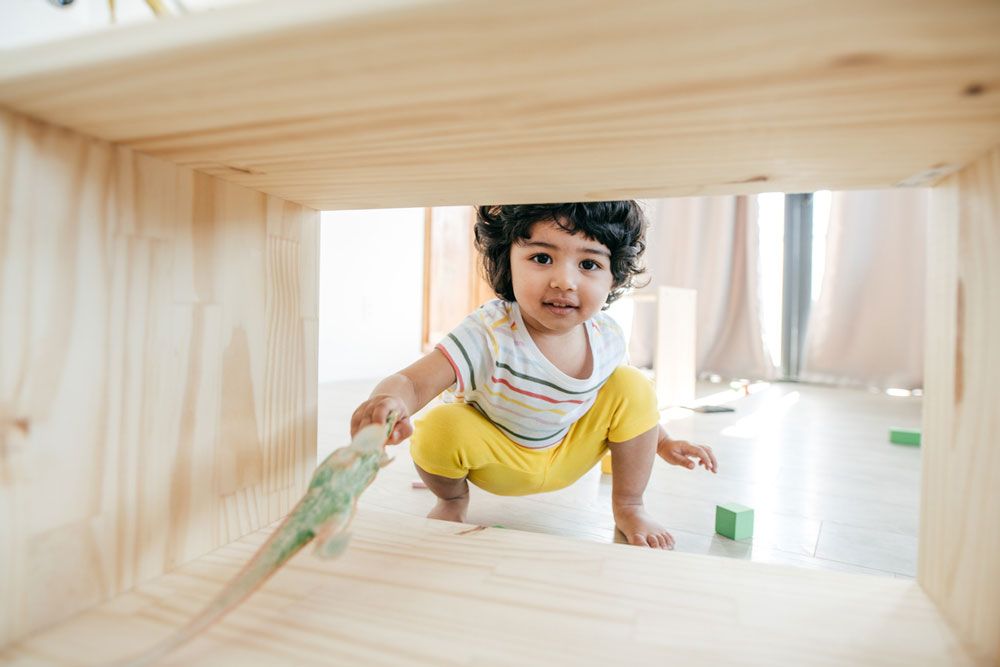 A curly-haired toddler in a striped shirt and yellow pants crouches and plays with a toy dinosaur and blocks.
