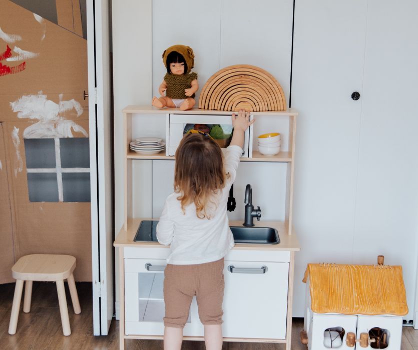 Toddler with back to camera reaches for a wooden rainbow on top of a play kitchen with a doll sitting nearby.