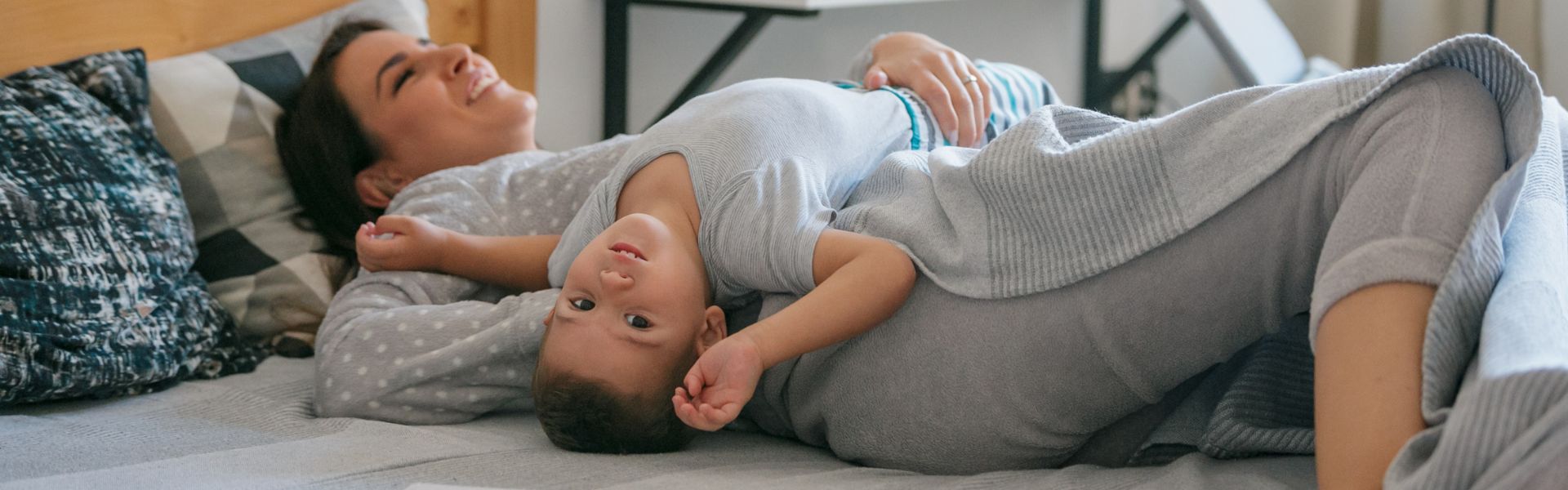 Smiling mother laying on a bed with a young child resting upside down on her chest.