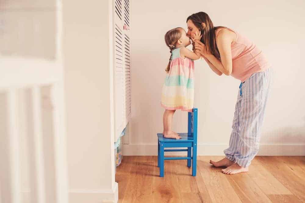 Toddler standing on a blue chair leans in to kiss a pregnant woman's nose.