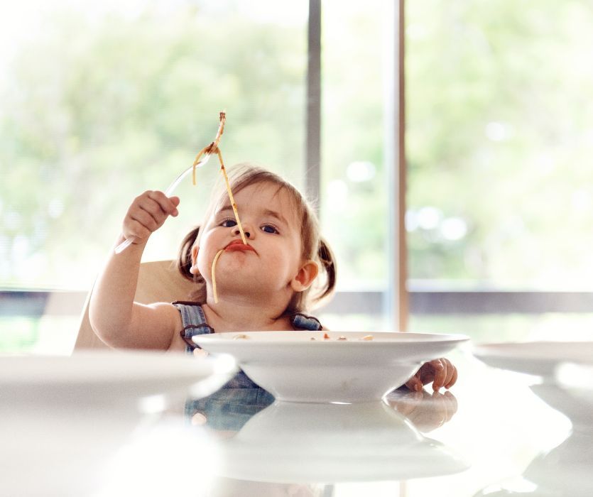 A toddler with a messy face eats spaghetti with a fork while sitting at a table.