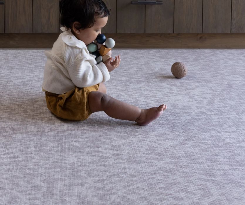 A baby sits on a grey patterned Totter & Tumble play mat playing with a colorful wooden toy.