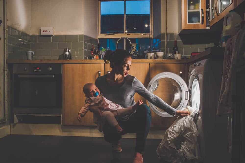 A woman holding a baby in a kitchen while crouched down to reach into an open, glowing front-load washing machine.