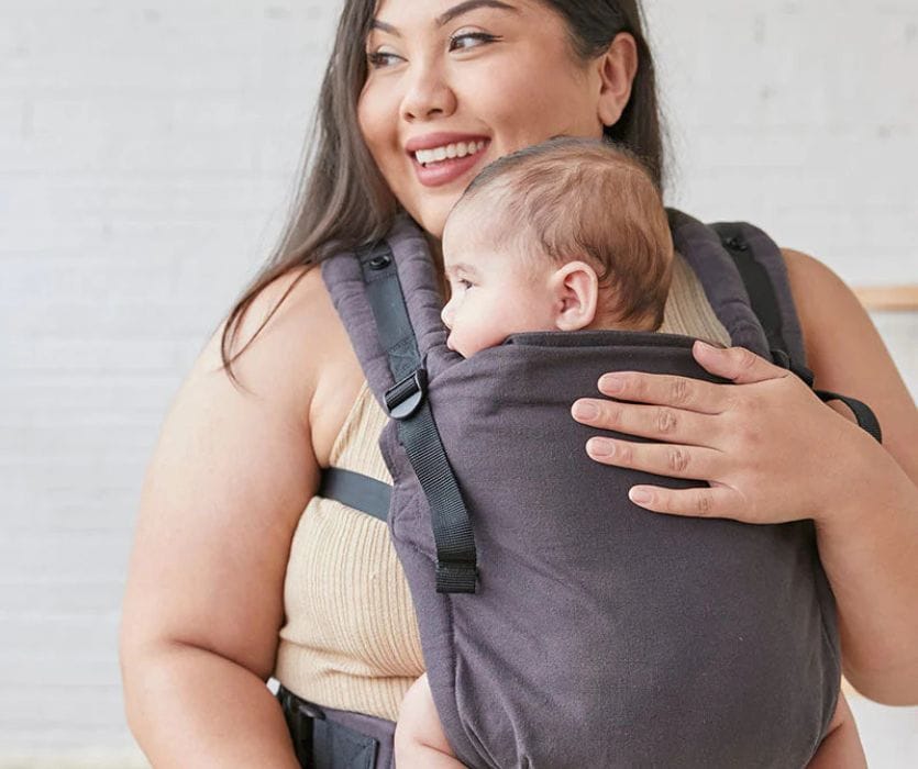 A smiling woman wearing a soft, dark gray Tula structured baby carrier with an infant inside.