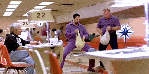 A man in a purple jumpsuit and a man in a purple polo shirt polish bowling balls with bags at a bowling alley.