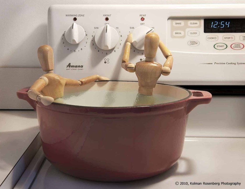 Two wooden artist mannequins sitting in a red pot of water on a stove, one turning a burner knob.