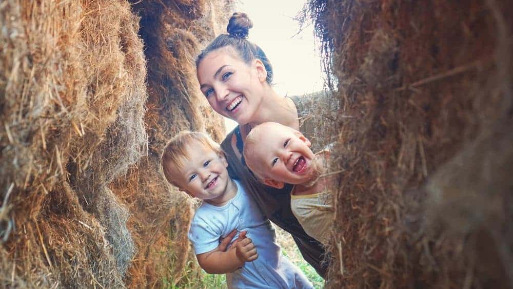 A smiling woman and two laughing young children peering through an opening between large hay bales.
