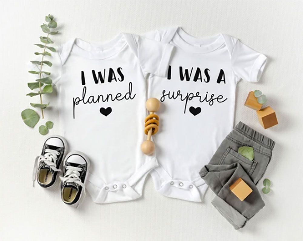 Two white baby onesies with text: I WAS planned and I WAS A surprise, next to tiny shoes, pants, and a wooden rattle.