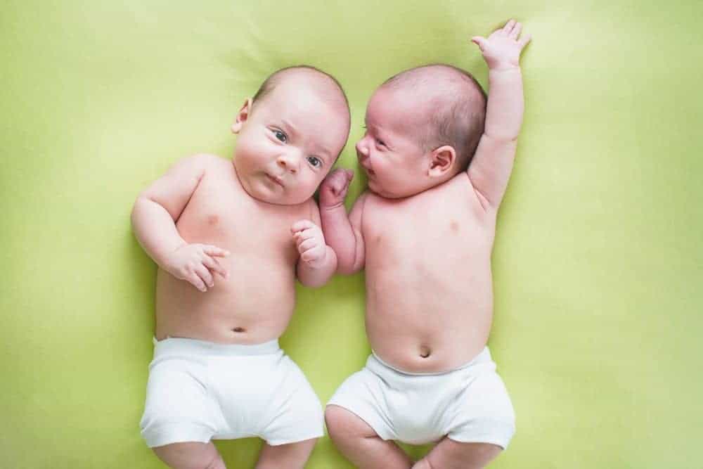 Top-down view of twin infants in white diapers lying side-by-side on a lime green background.