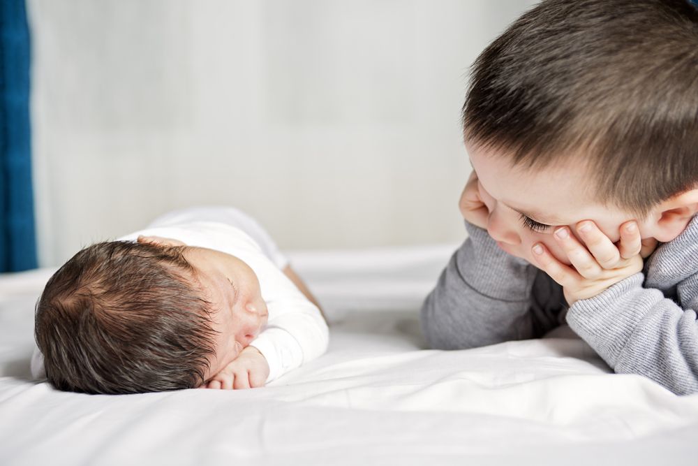 A young boy rests his head on his hands while watching a newborn baby sleep on a white bed.