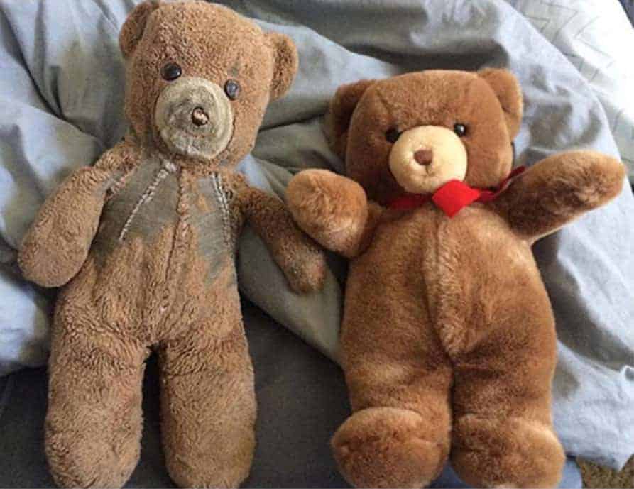 A well-worn brown teddy bear with visible repairs next to a new version of the same teddy bear with a red ribbon.